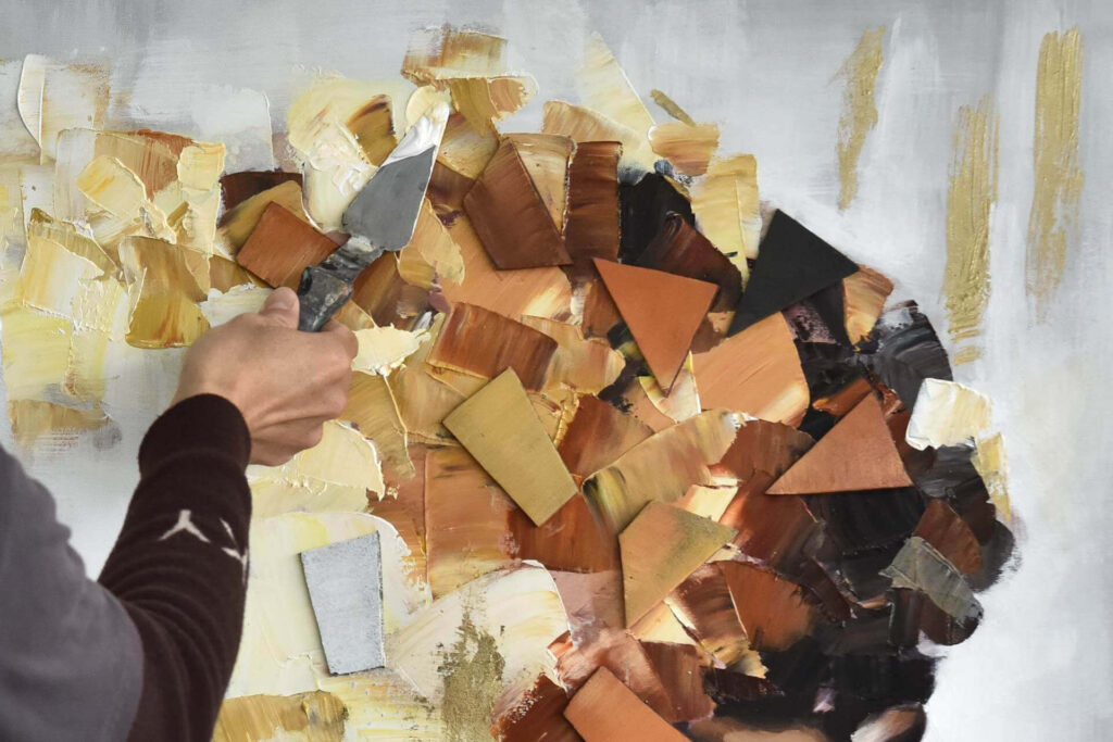 An artist using a palette knife to apply thick, 3D layers of paint to a modern abstract portrait with gold and brown tones.